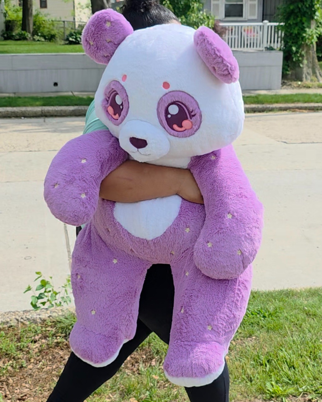 A person outdoors holding a large plush panda toy. The panda is white on the face and chest, with purple ears, purple arms, and purple legs, decorated with small pink and yellow embroidered details.